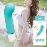 A turquoise VeDO 'Gee' Mini G-Spot Rechargeable Waterproof Vibrator, crafted from medical-grade silicone, is displayed alongside its packaging in the foreground. In the background, an undressed individual is posed outdoors with a scenic view. The product box emphasizes its 10 modes of vibration and brand details for optimal G-spot stimulation.